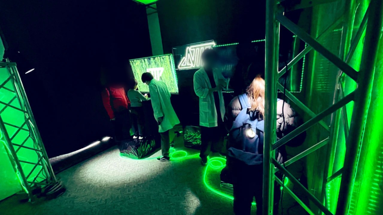 緑のネオンで照らされた NEO TOKYO PUNKS の体験会場。白衣のスタッフの案内でモニターの謎解きに挑む参加者(Neo Tokyo Punks 地方自治体 イベント制作)